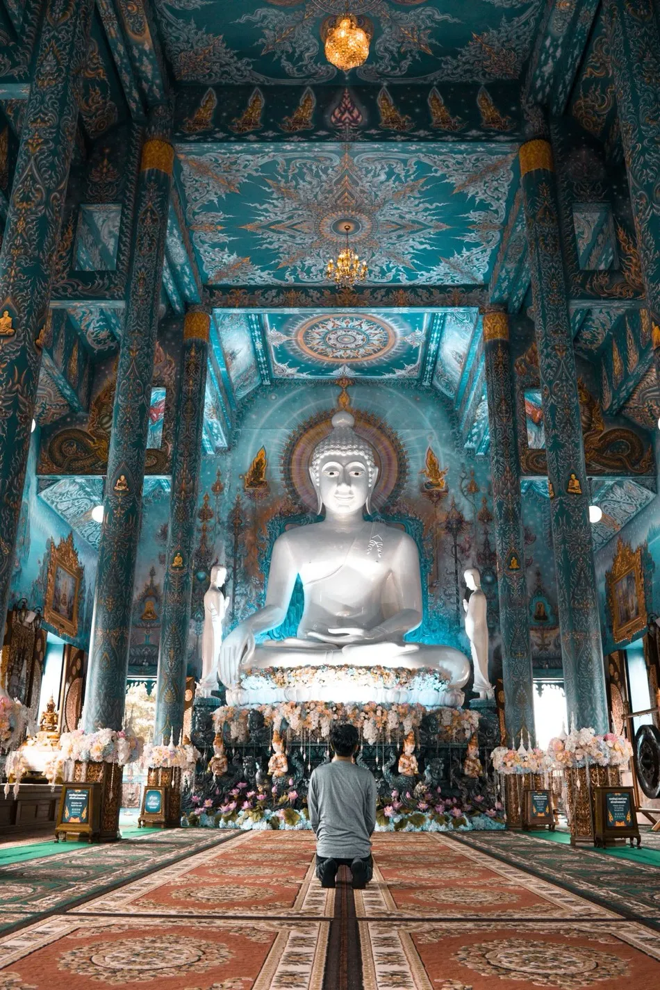A lone worshipper kneeling before a large white Buddha in an intricately decorated hall